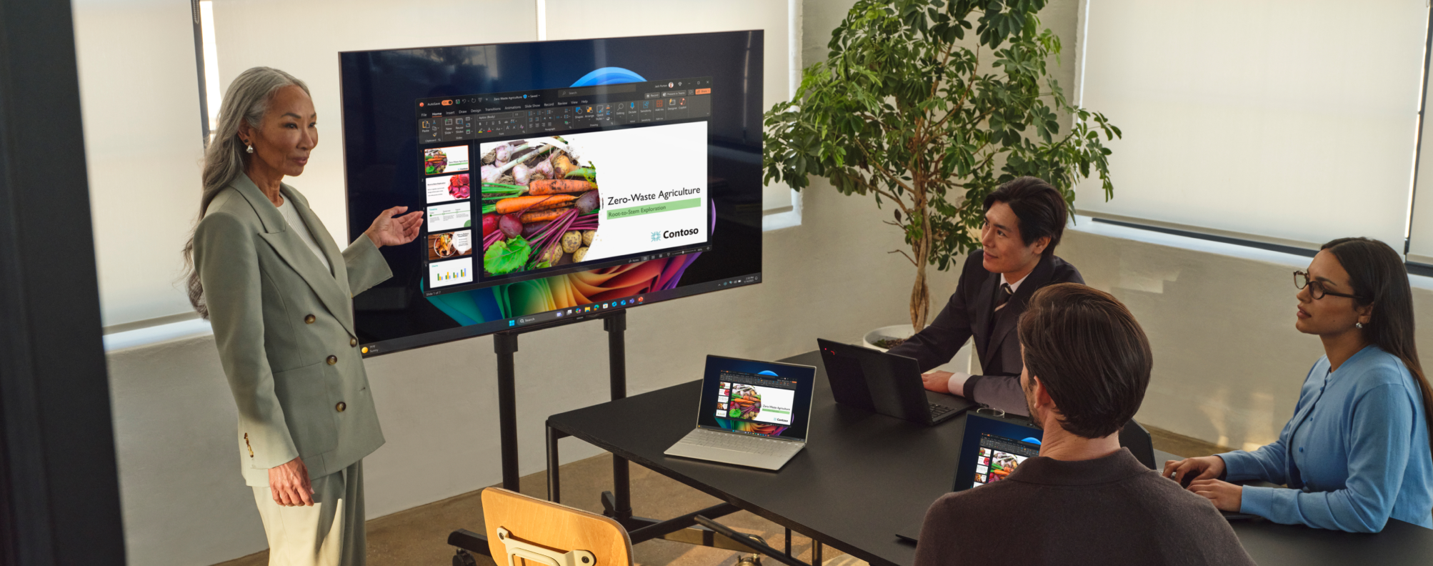 Four people in a conference room, three sitting at a table watching the fourth present at a large display screen.