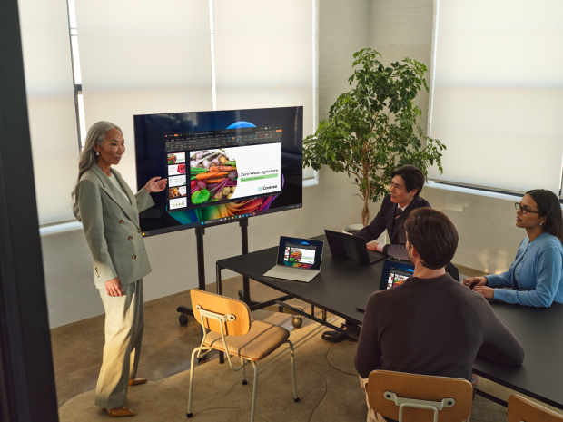 Four people in a conference room, three sitting at a table watching the fourth present at a large display screen.